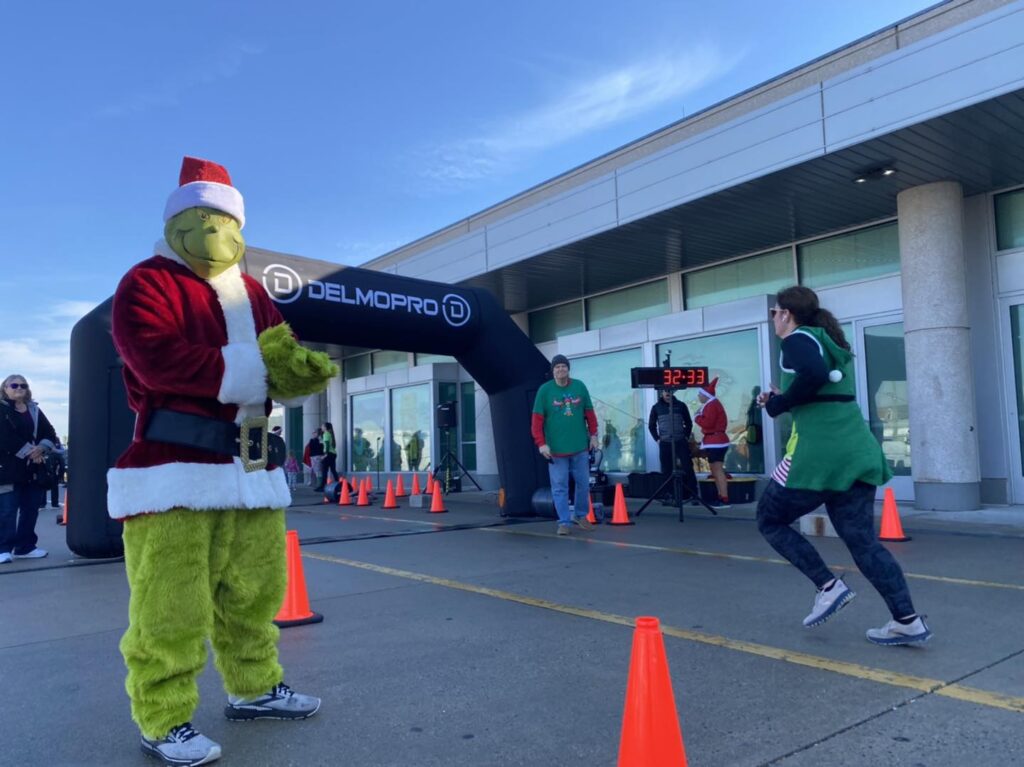 Pinch the ‘GrYnch’ Boardwalk 5K Run with Runners racing towards finish line where person dressed in Grinch costume is cheering racers on in the Wildwoods NJ boardwalk